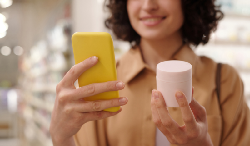 Female consumer examining a small pink pot of cream holding her smart phone
